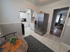 A modern kitchen with stainless steel appliances, a dining table with a leaf-patterned centerpiece, and a hallway leading to a living area. - Cozy Allen Park Bungalow (Allen Park)