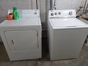 Two white washing machines side by side in a utility room with a concrete floor and a white brick wall. - Cozy Allen Park Bungalow (Allen Park)