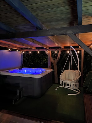Un jacuzzi bajo una pérgola de madera con luces colgantes, rodeado de follaje.