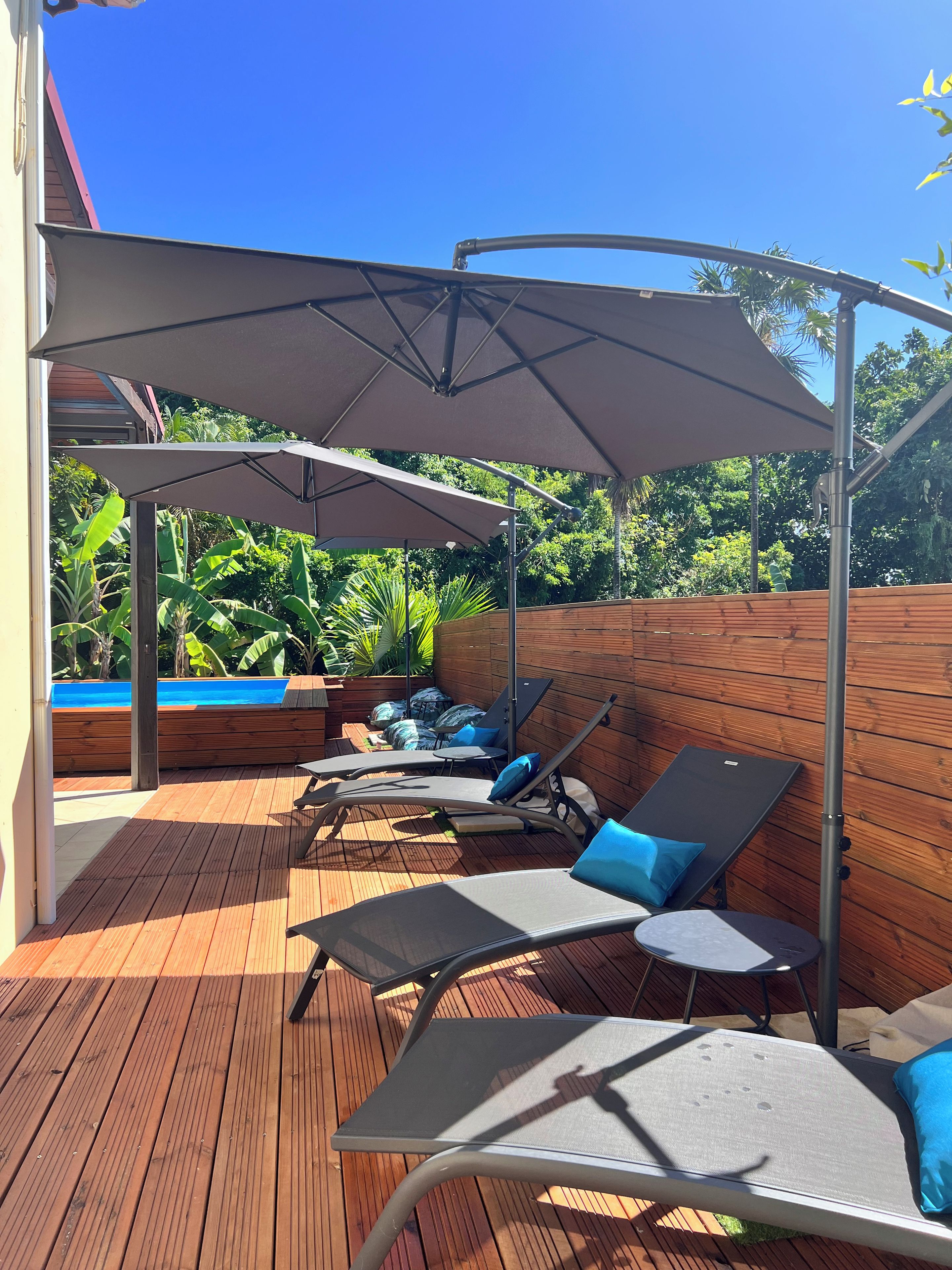 A wooden deck with lounge chairs and umbrellas, a pool, and lush greenery in the background.