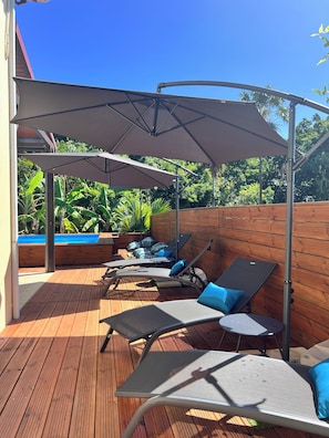 A wooden deck with lounge chairs and umbrellas, a pool, and lush greenery in the background.