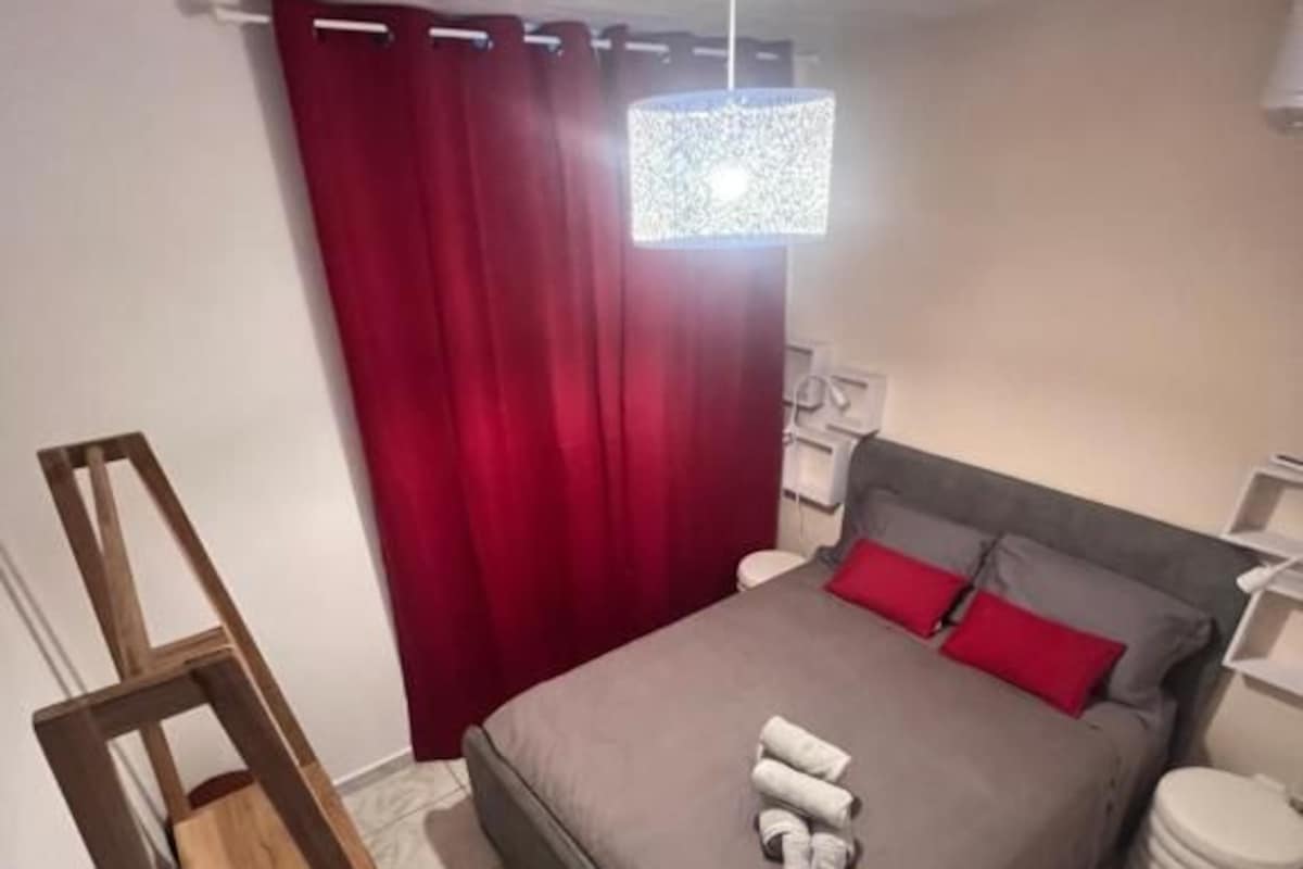 A bedroom with a bed, a wooden ladder, a red curtain, a white bedside table, and a white lamp.