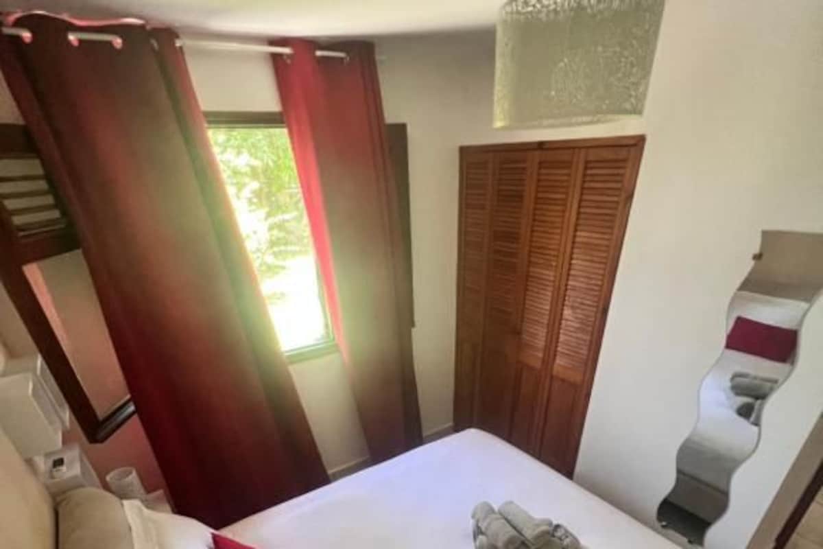 A hotel room with a bed, a sofa, a window with curtains, a wooden door, and a wall-mounted light fixture.