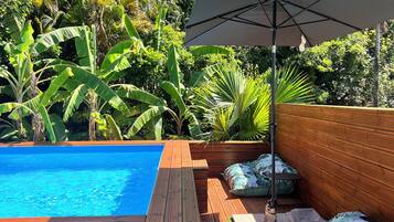 Una terraza de madera con piscina, rodeada de abundante vegetación y un gran paraguas.
