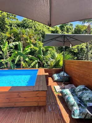 Una terraza de madera con piscina, rodeada de abundante vegetación y un gran paraguas.