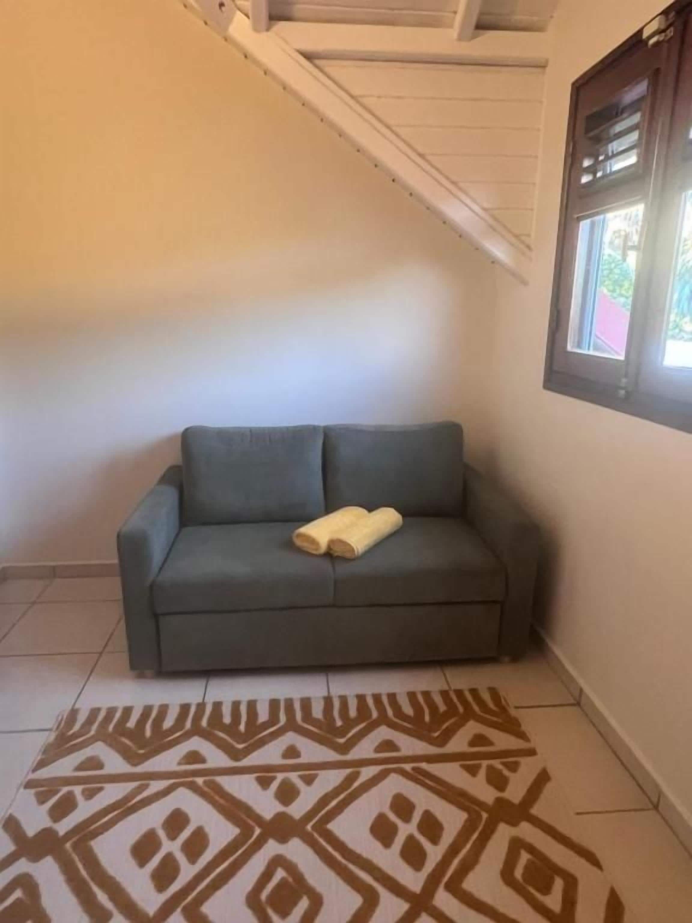 A grey sofa with a yellow towel, a patterned rug, and a window with blinds.