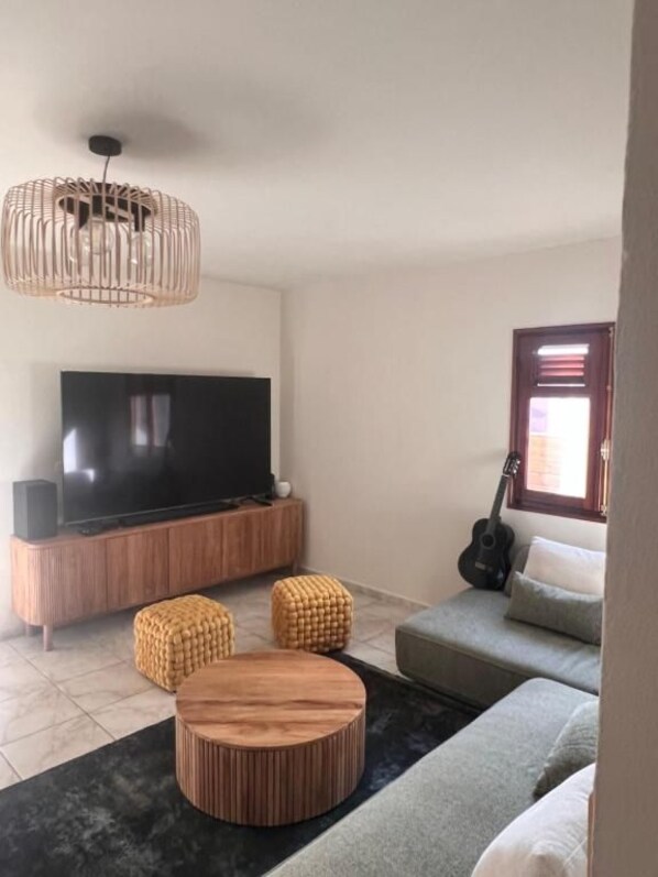 A living room with a flat-screen TV, a wooden coffee table, a grey sofa, and a guitar leaning against the wall.