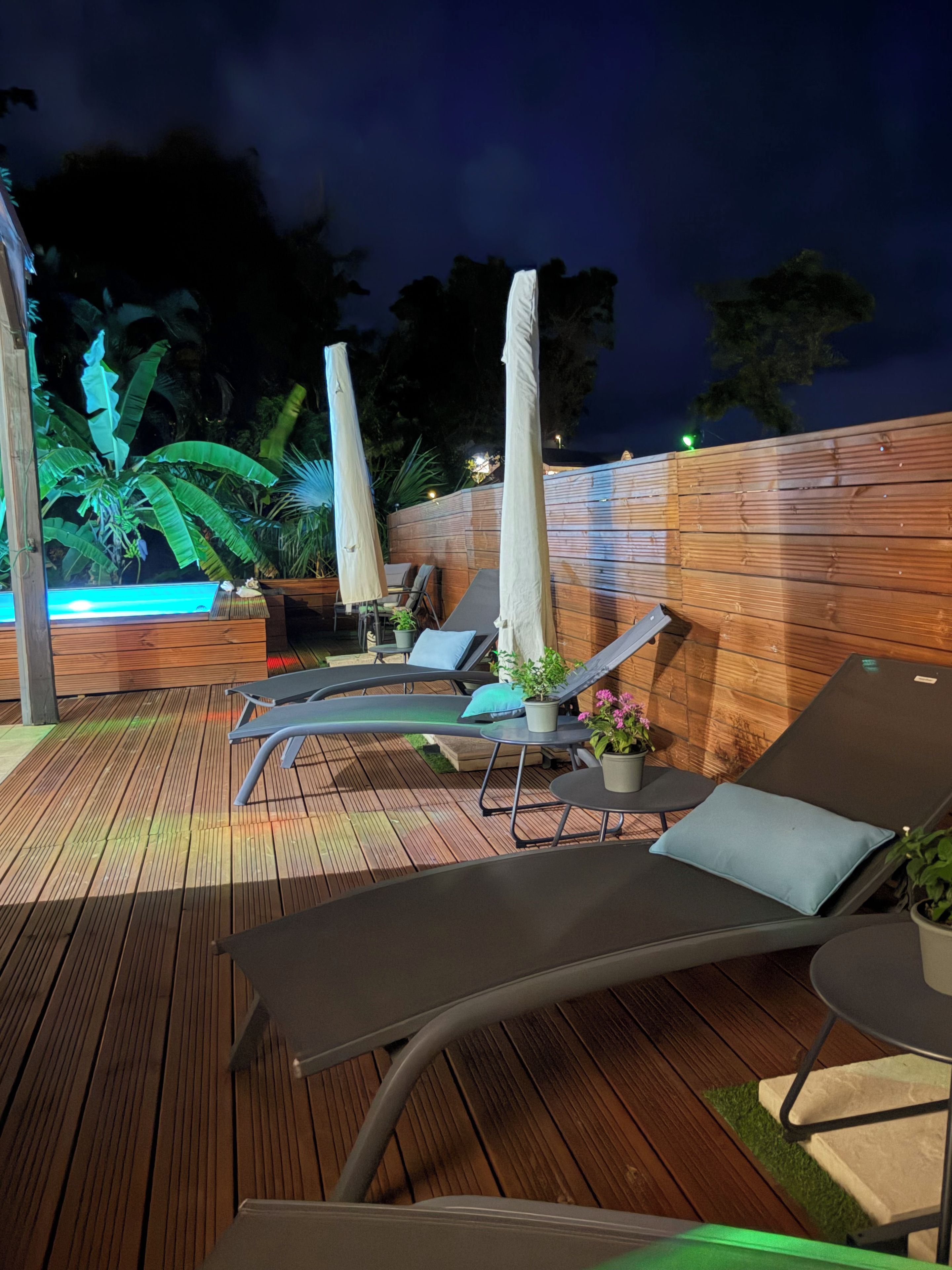 Une terrasse extérieure avec des chaises longues et des parasols, entourée d’une clôture en bois et agrémentée de plantes en pot.