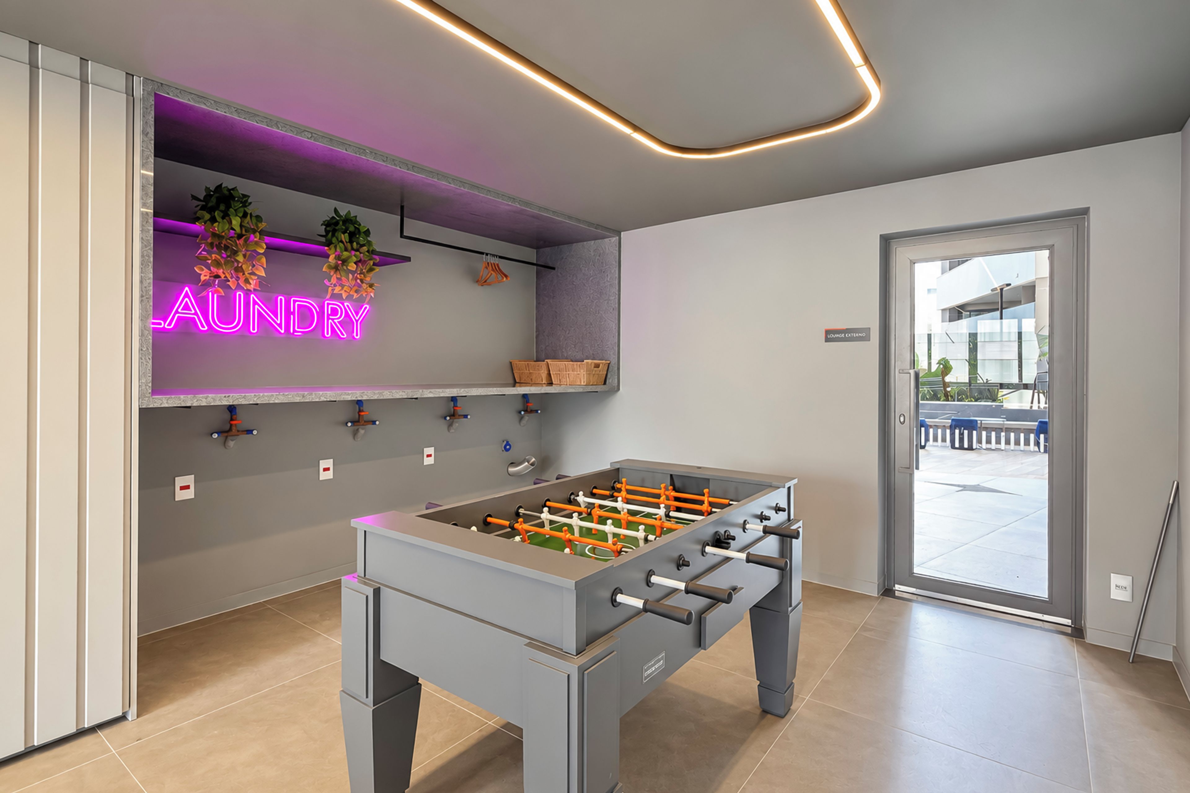 A modern room with a foosball table, a neon sign reading 'AUNDRY', and a door leading to an outdoor area.