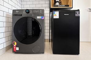 Una unidad compacta de lavadora y secadora con pantalla digital, un refrigerador compacto y un estante de madera con una cafetera.