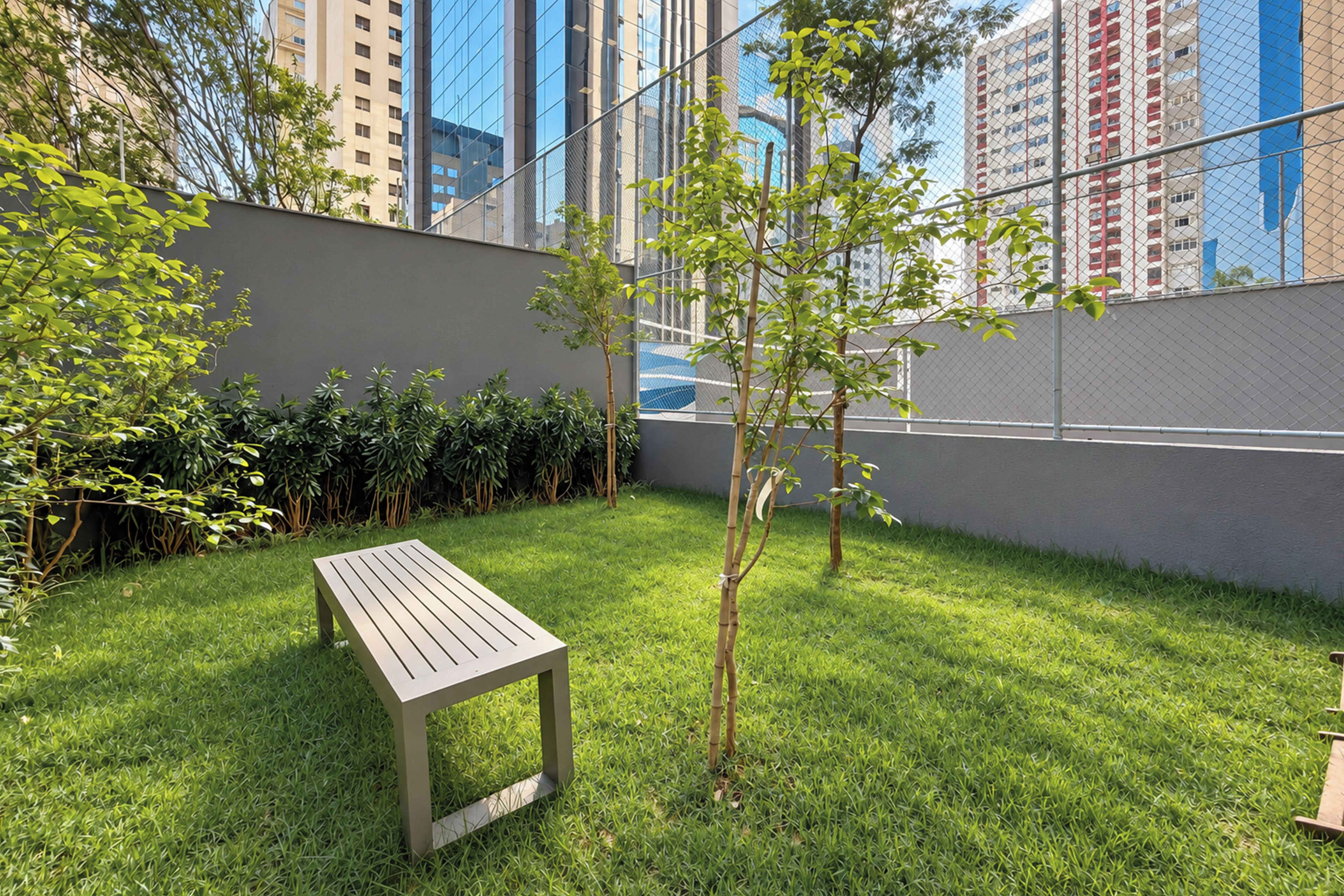 A grassy outdoor area with a wooden bench, a small tree, and a chain-link fence.