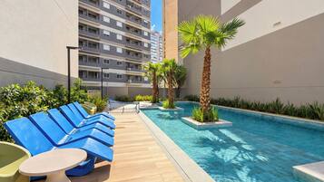 A rooftop pool area with blue lounge chairs and a small table, surrounded by greenery and a tall building in the background.
