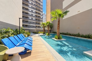 Un área de piscina en la azotea con sillas reclinables azules y una pequeña mesa, rodeada de vegetación y un edificio alto al fondo.