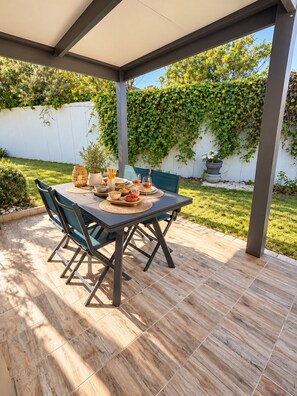 Un patio al aire libre con una mesa lista para comer, rodeado de sillas y un área cubierta con vistas a un jardín y una pared blanca.