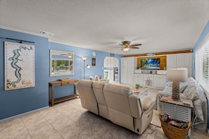 A living room with a beige sofa, a dining area with a TV, and a kitchen in the background. - Fisherman's Reel Retreat: Unit 2 (Main home only) (Satsuma)