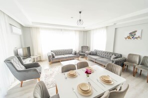 Ein geräumiges Wohnzimmer mit einem grauen Ecksofa, einem Esstisch für vier Personen und einem Fernseher links.