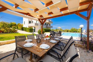 An outdoor dining area with a wooden table and chairs, a pool in the background, and a clear sky. - Villa Diamantis - Six Bedroom Villa, Sleeps 12 (Rethymno)