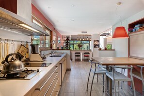 A modern kitchen with a gas stove, kettle, and various utensils. There is a dining area with a table and chairs, and a window providing a view of the outdoors. - Villa 'Terre de Lumières' with View of Mount Néron in a Natural Setting (Quaix-en-Chartreuse)