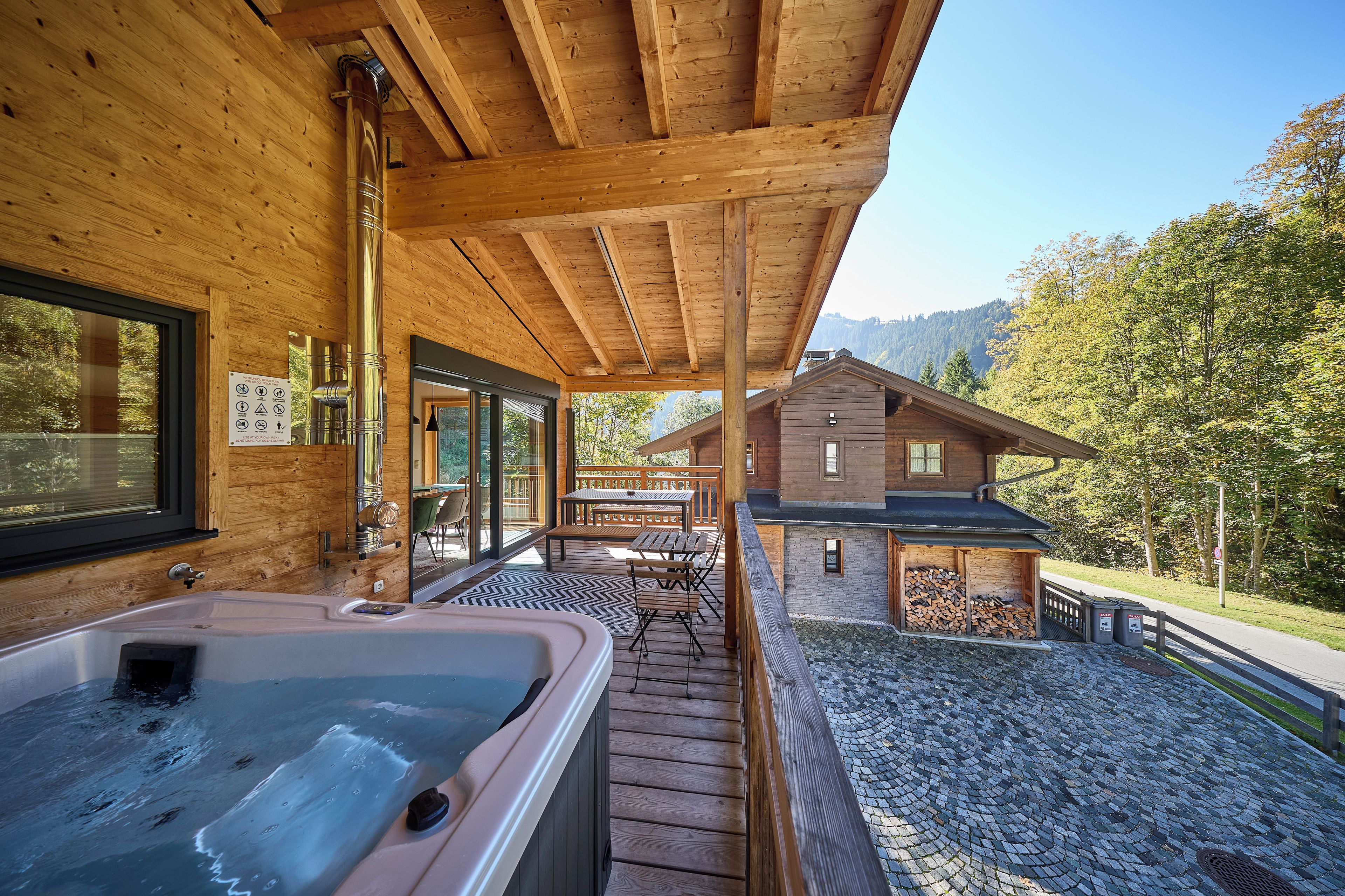 A wooden cabin with a hot tub, a deck, and a view of the surrounding forest.