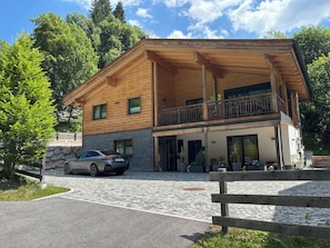 A two-story wooden house with a balcony, a stone wall, and a parked car. - Chalets Glemmtal - Chalet Bergblick (Viehhofen)
