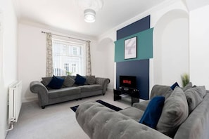 A modern living room with a grey sofa, a flat-screen TV, and a large window with curtains. - Spacious and stylish 3 bedroom home,fully equipped kitchen and relaxing bedrooms (Jarrow)