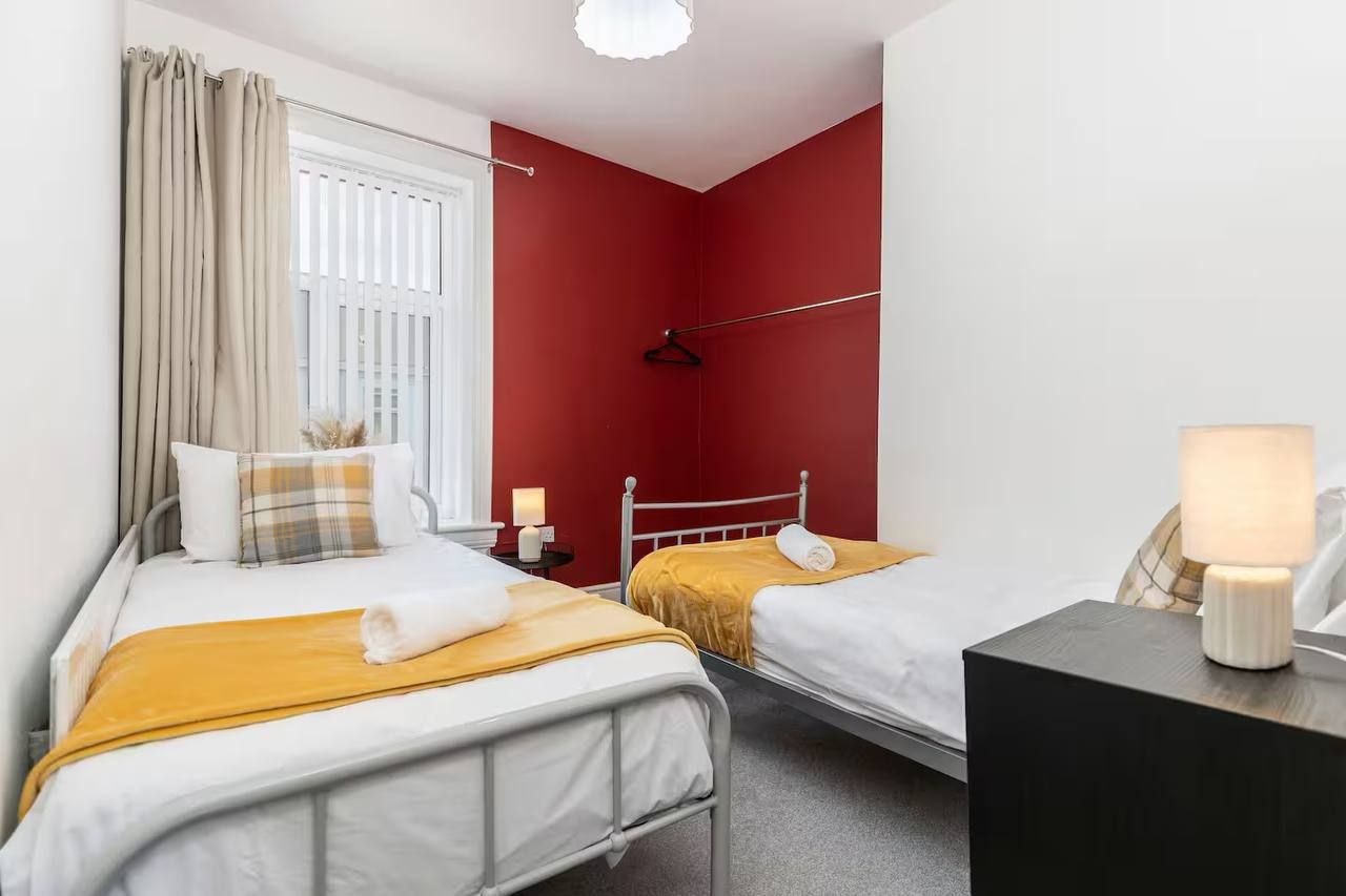Habitación de hotel con dos camas, una mesita de noche con lámpara, una ventana con cortinas y una pared de acento roja.