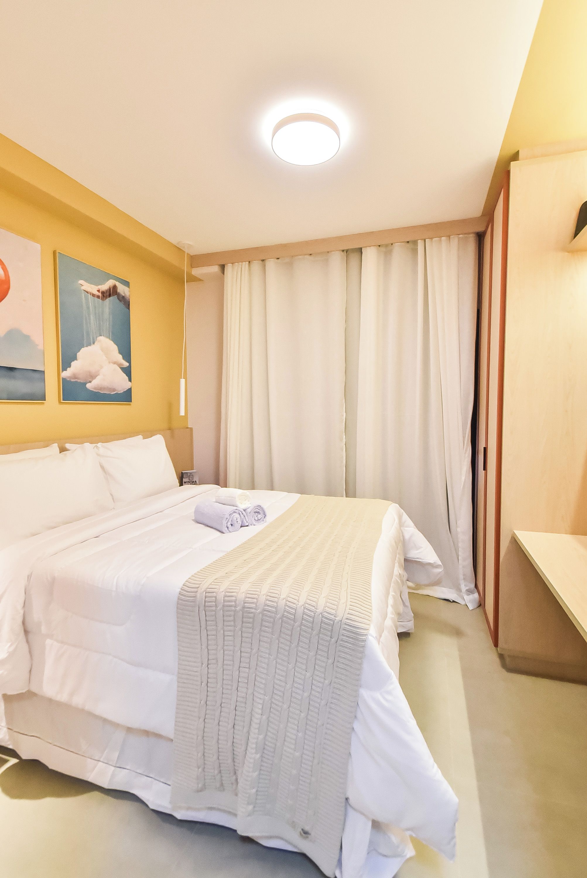 A hotel room with a bed, a desk, a nightstand, a wall-mounted lamp, and a window with curtains.