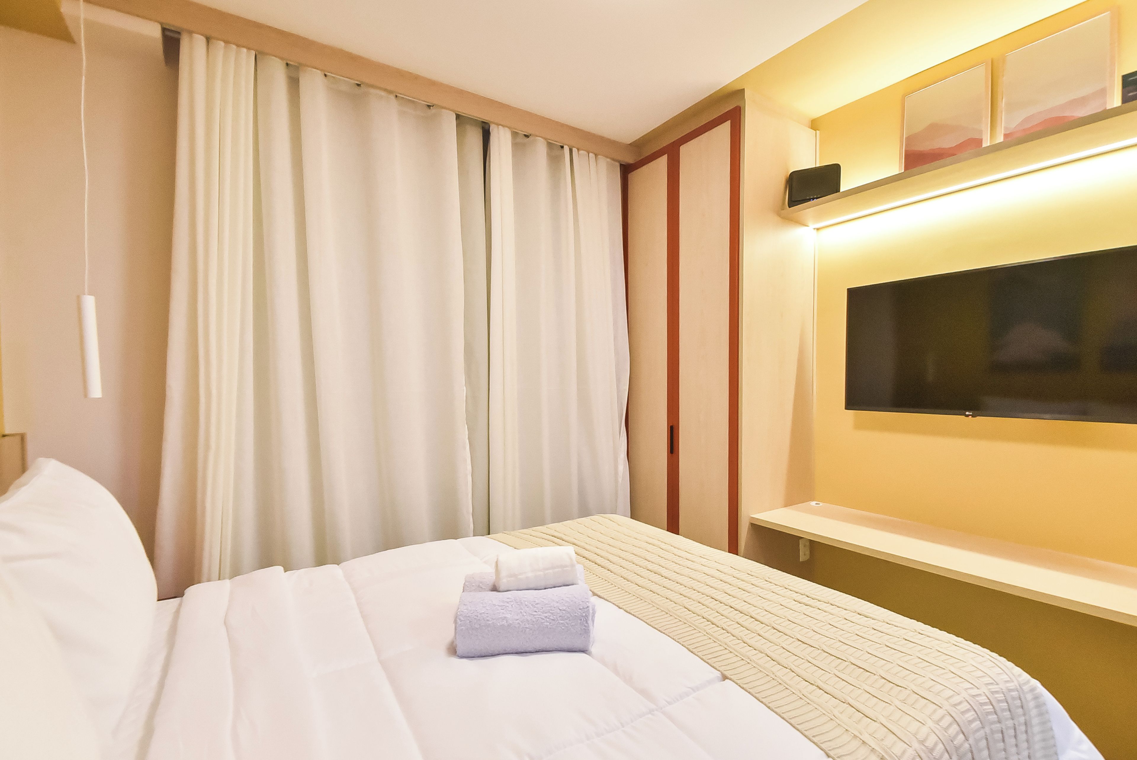 A hotel room with a bed, a flat-screen TV mounted on the wall, and a window with curtains.