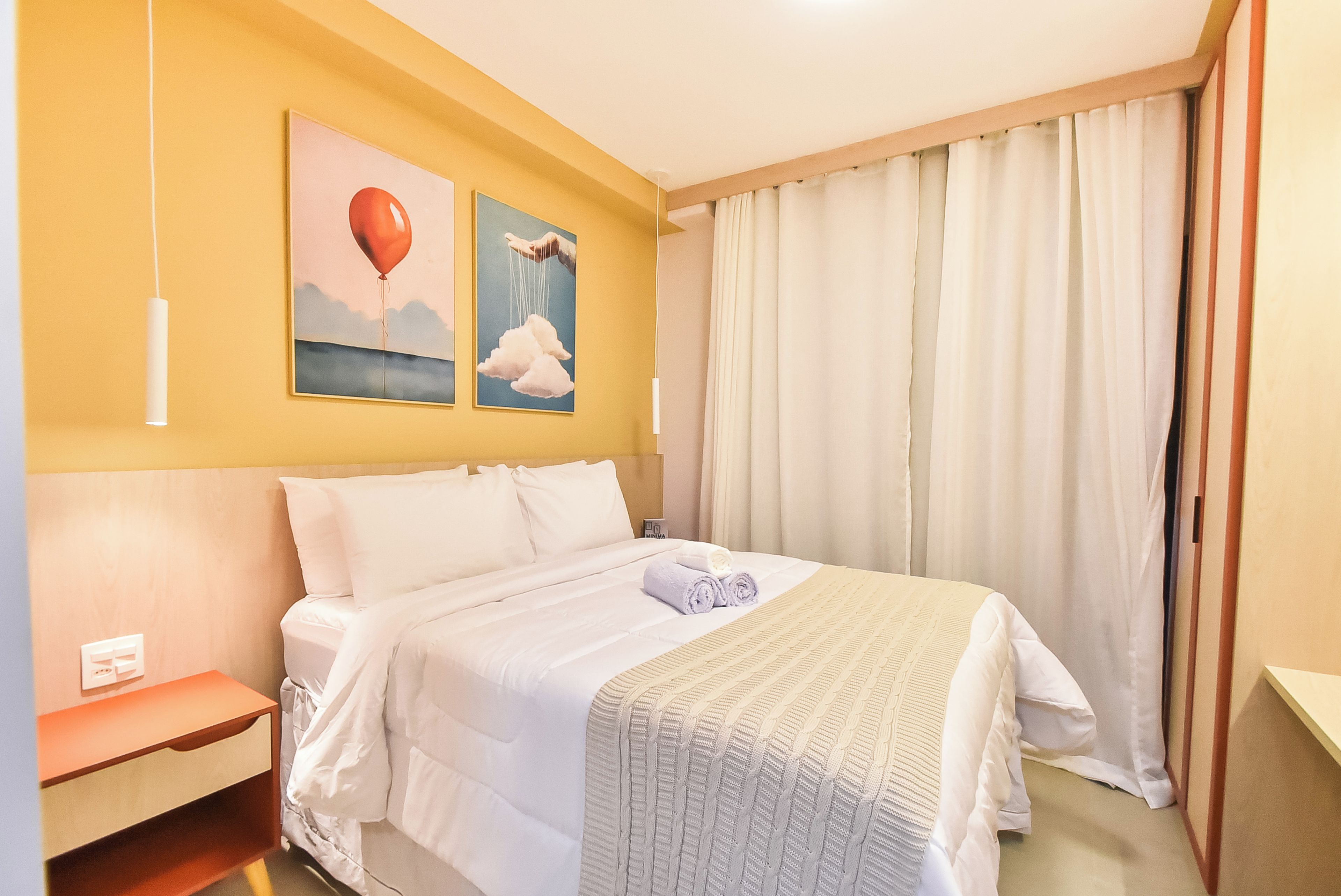 A hotel room with a bed, a nightstand, a painting of a hot air balloon, and a window with curtains.