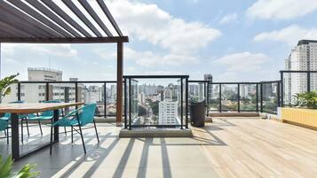 An outdoor terrace with a wooden floor, a table with chairs, a glass partition, and a view of a cityscape.