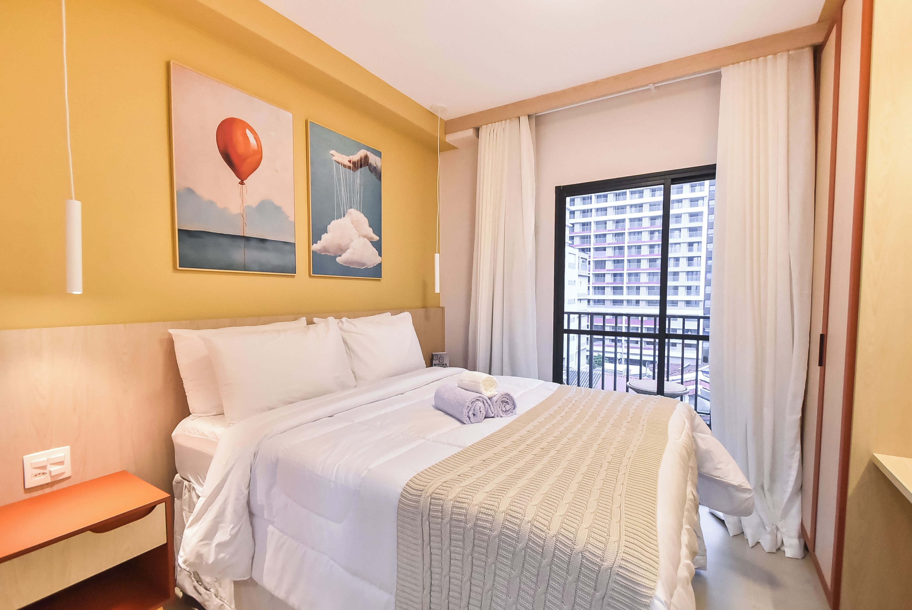 A hotel room with a bed, a bedside table, a window with curtains, and two wall art pieces.