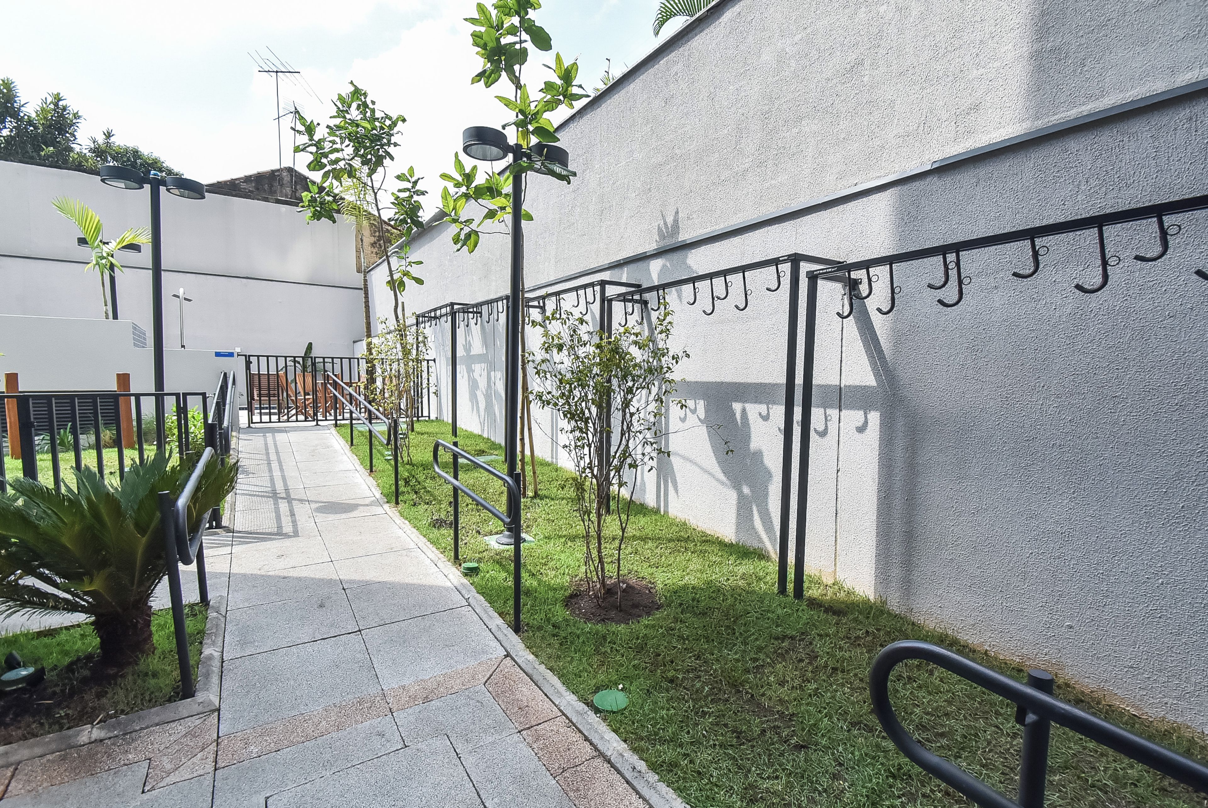 A paved pathway leading to a building with a white exterior wall, a metal fence with hooks, and a small tree.