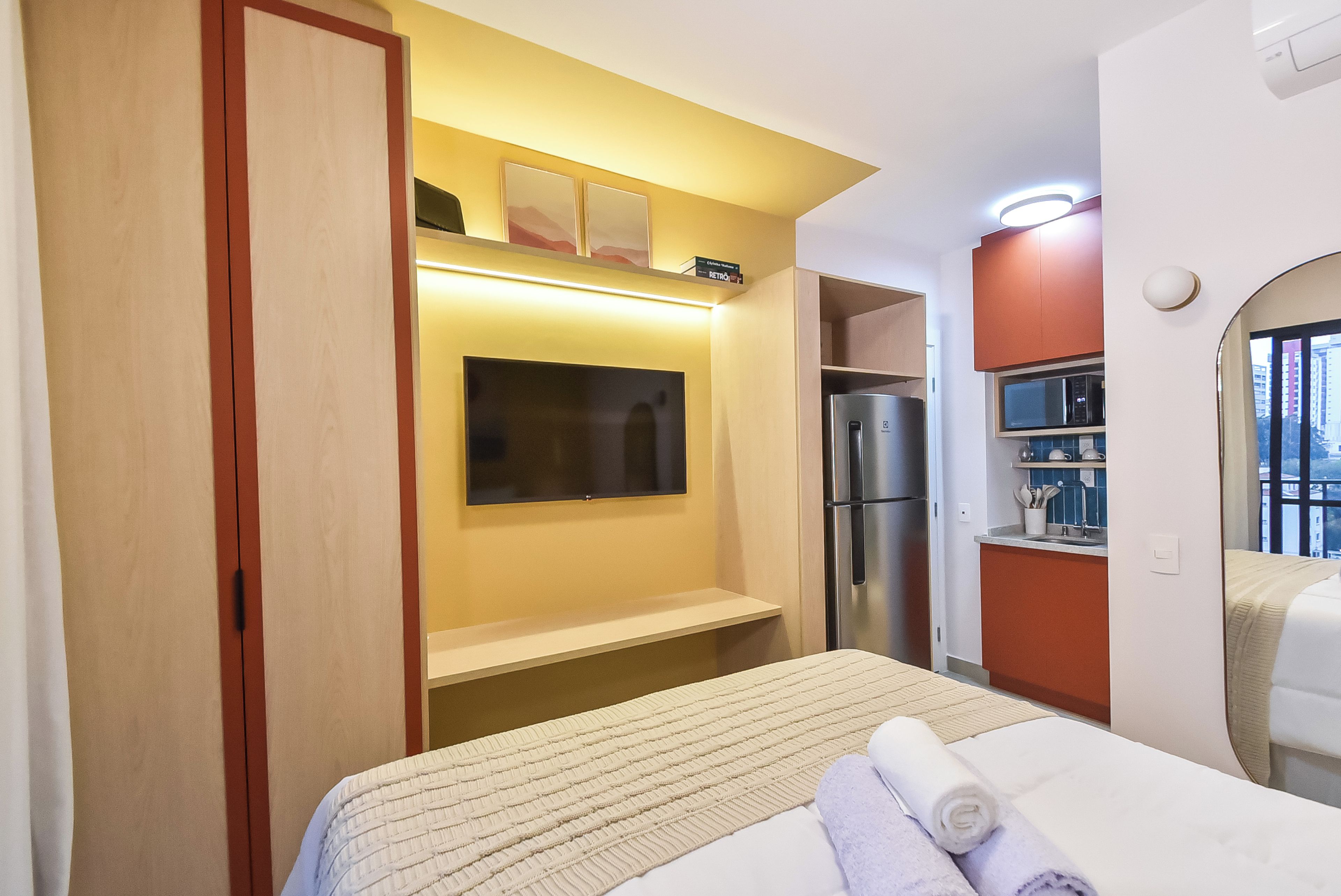 A hotel room with a bed, a flat-screen TV mounted on the wall, a refrigerator, and a microwave.