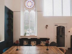 A dining area with a wooden table, chairs, a bench with floral cushions, a stained glass window, and a door with a glass panel. - Chapel of Arts (Criccieth)