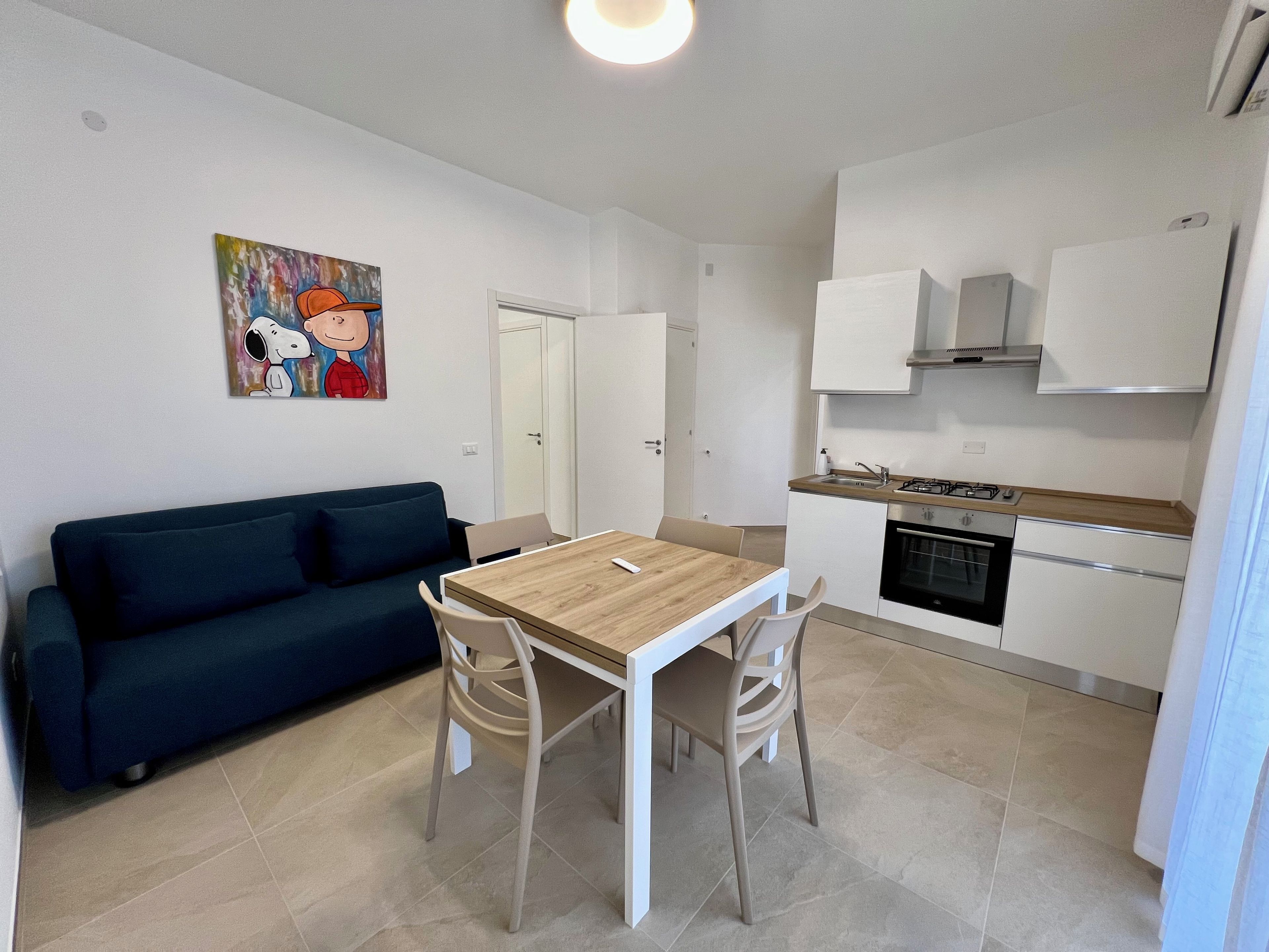 A compact living space with a kitchenette, a dining area with a table and chairs, a dark-colored sofa, and a wall-mounted painting.