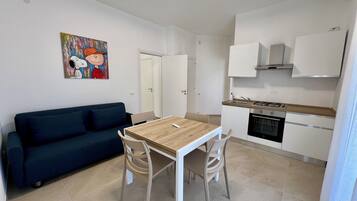 A compact living space with a kitchenette, a dining area with a table and chairs, a dark-colored sofa, and a wall-mounted painting.