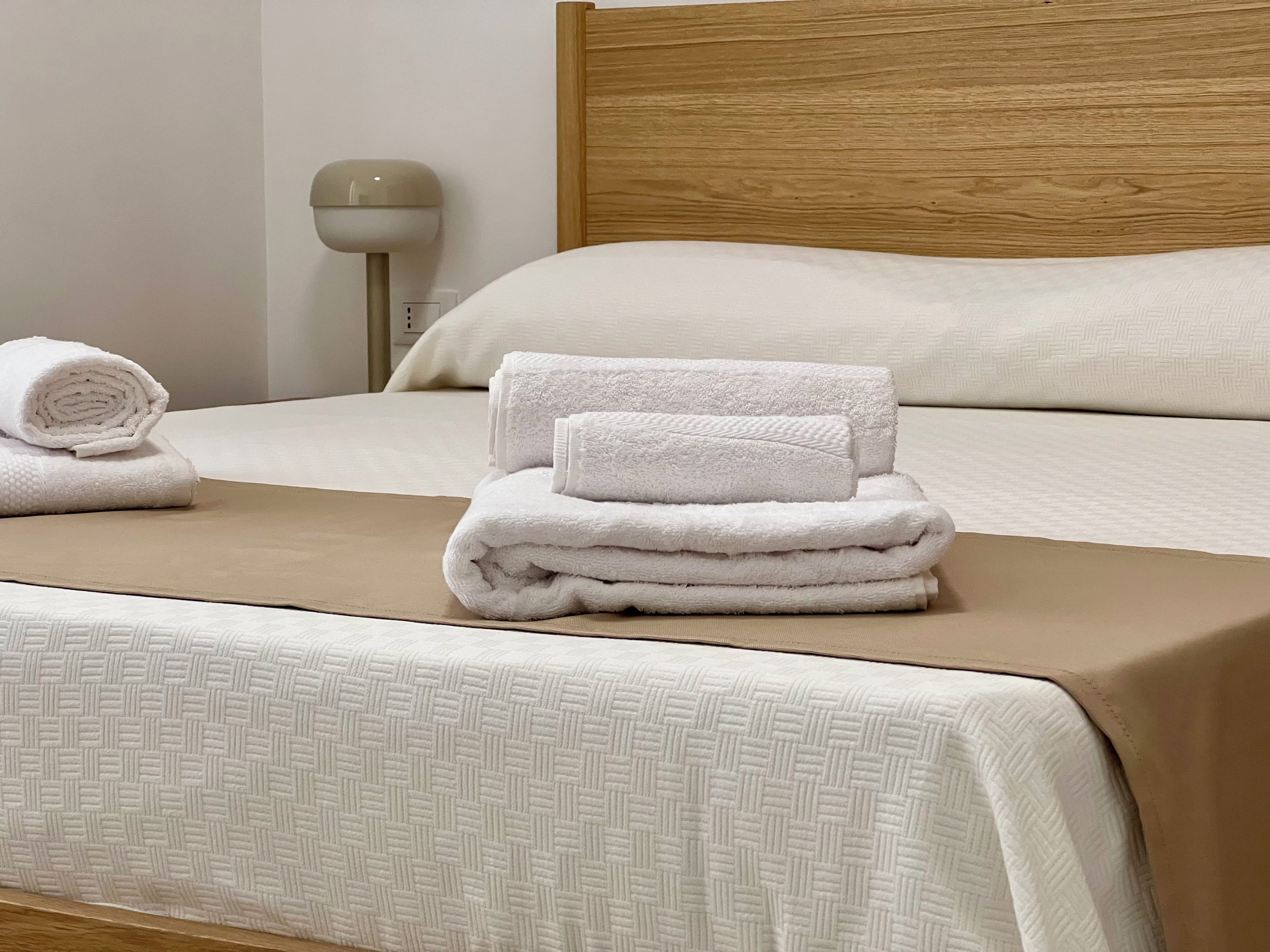 A neatly made bed with a beige headboard, a rolled towel, and a stack of folded towels on the bed.