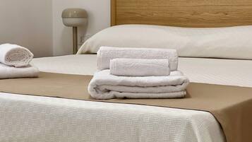 A neatly made bed with a beige headboard, a rolled towel, and a stack of folded towels on the bed.