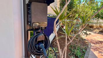An outdoor electrical charging station attached to a building, surrounded by greenery and a paved pathway.
