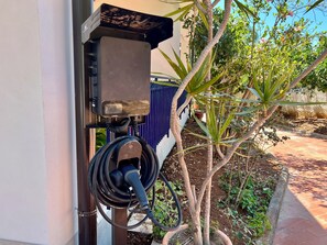 Une borne de recharge électrique extérieure, fixée à un bâtiment, entourée de verdure et bordée d’un chemin pavé.