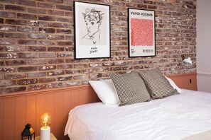 Une chambre d’hôtel avec un mur en briques, un lit avec une literie blanche, deux coussins à motifs et deux œuvres d’art encadrées.