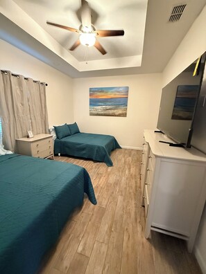 Une chambre avec deux lits, un ventilateur de plafond, un tableau et un téléviseur posé sur une commode.