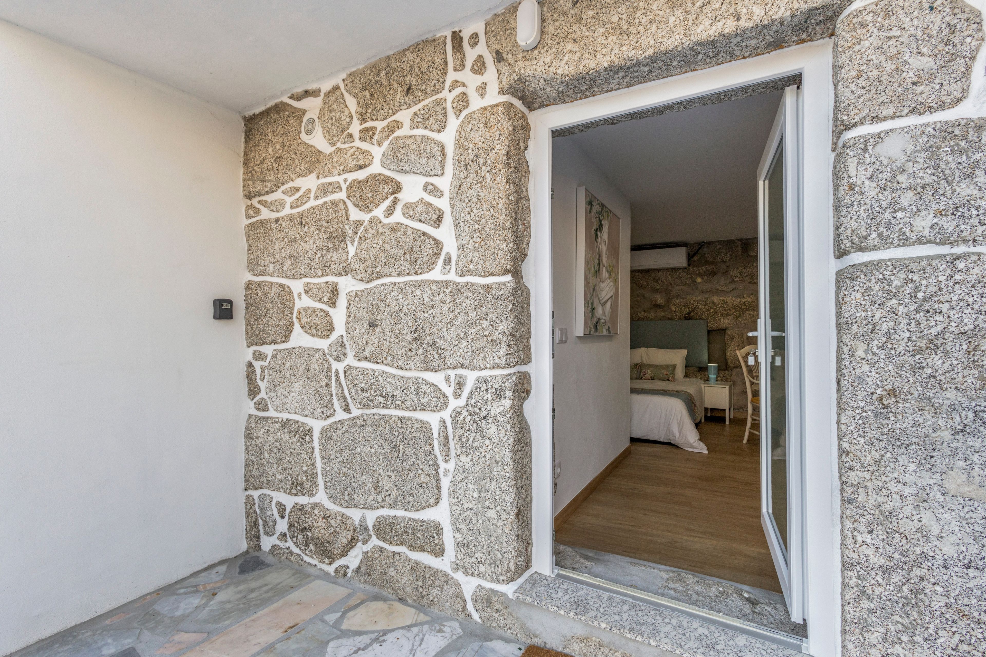 A stone wall with a doorway leading to a bedroom with a bed and a nightstand.