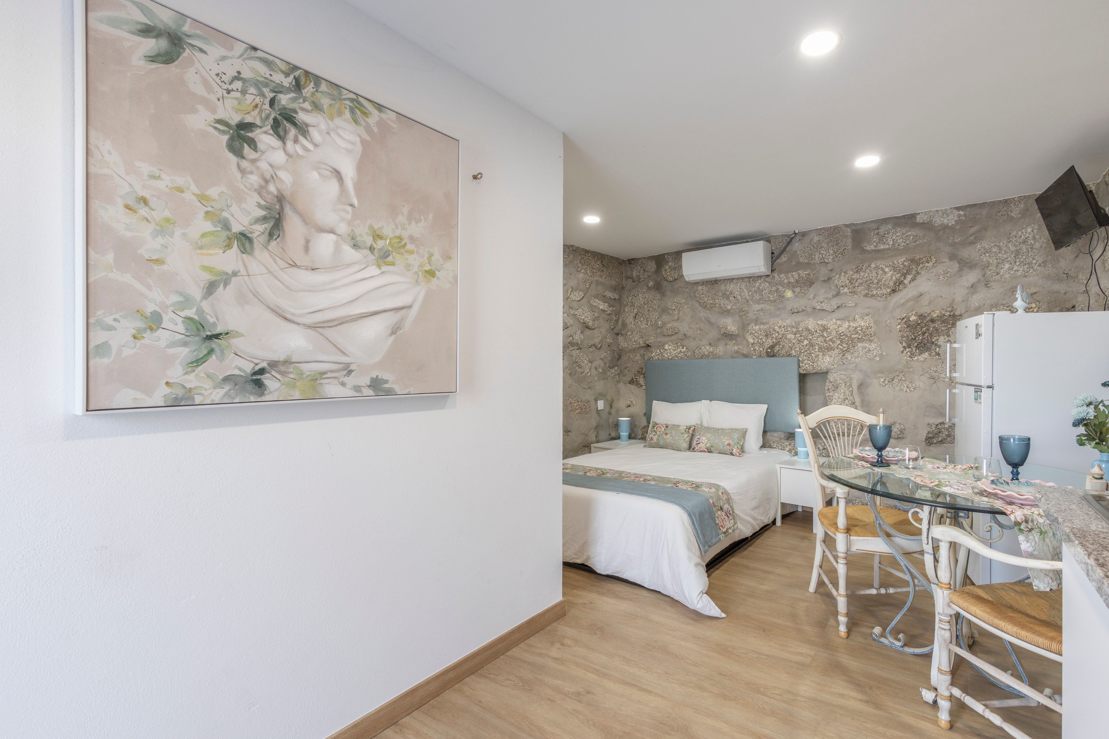 A room with a bed, a table, and a chair. There is a painting on the wall.