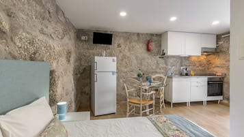 Habitación compacta con cama, una kitchenette pequeña, zona de comedor con mesa y sillas, y un televisor montado en la pared.