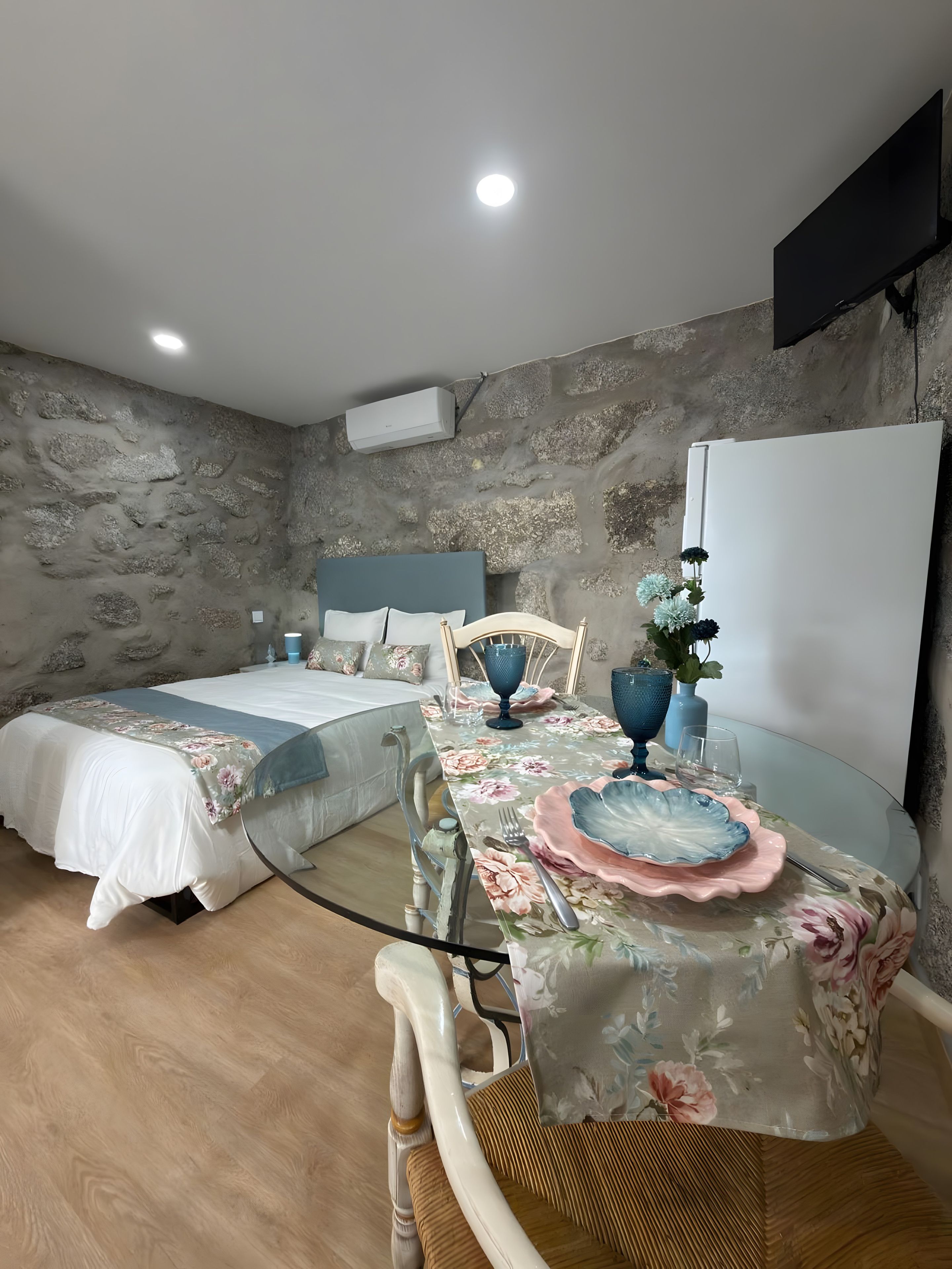 A bedroom with a bed, a table with a floral tablecloth, and a stone wall.