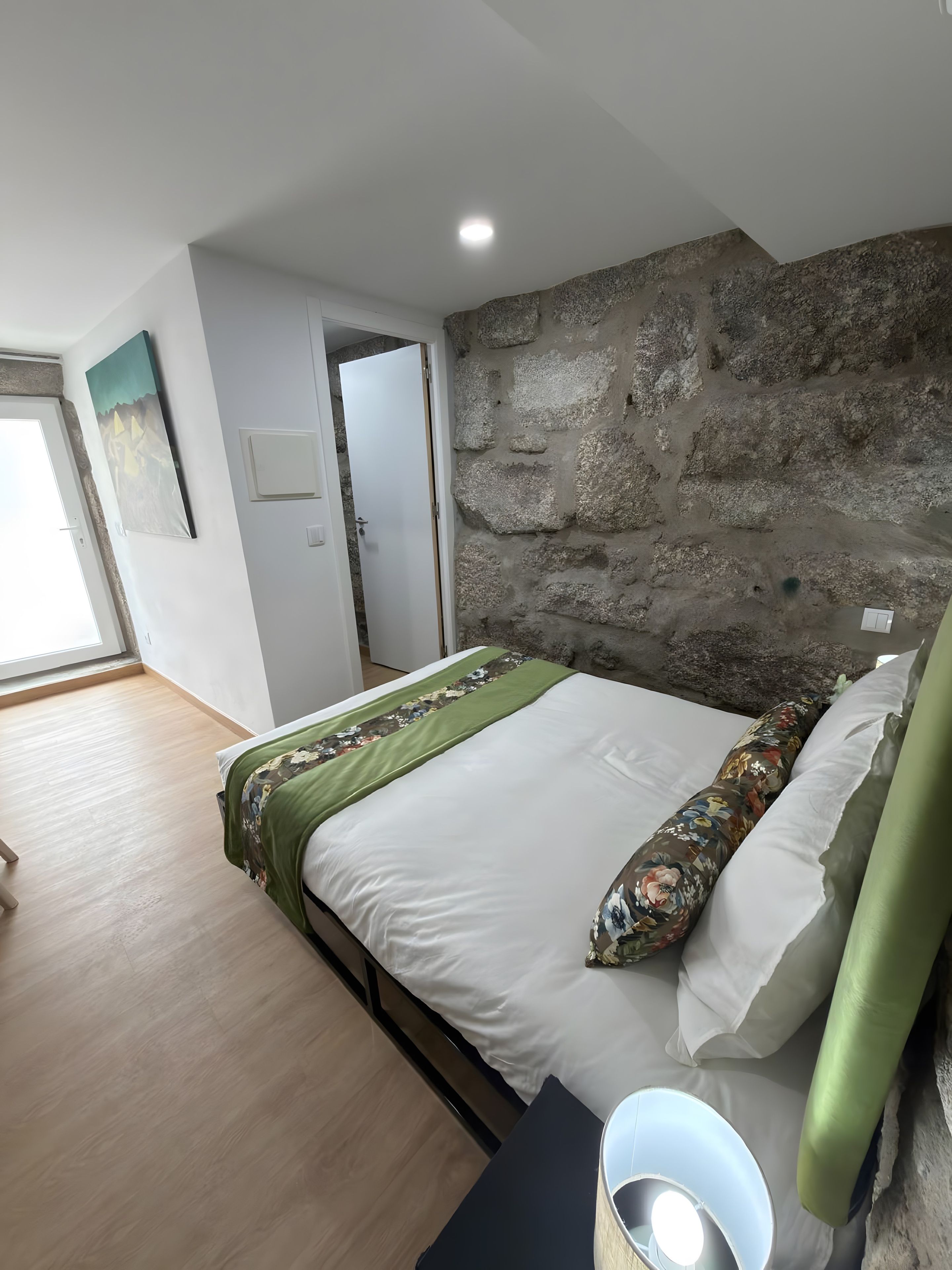 A bedroom with a stone wall, a bed with white bedding, a green throw, and a nightstand with a lamp.