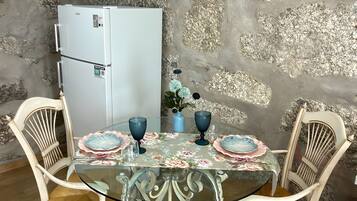 A dining area with a glass table, two chairs, a small refrigerator, and a mounted television.