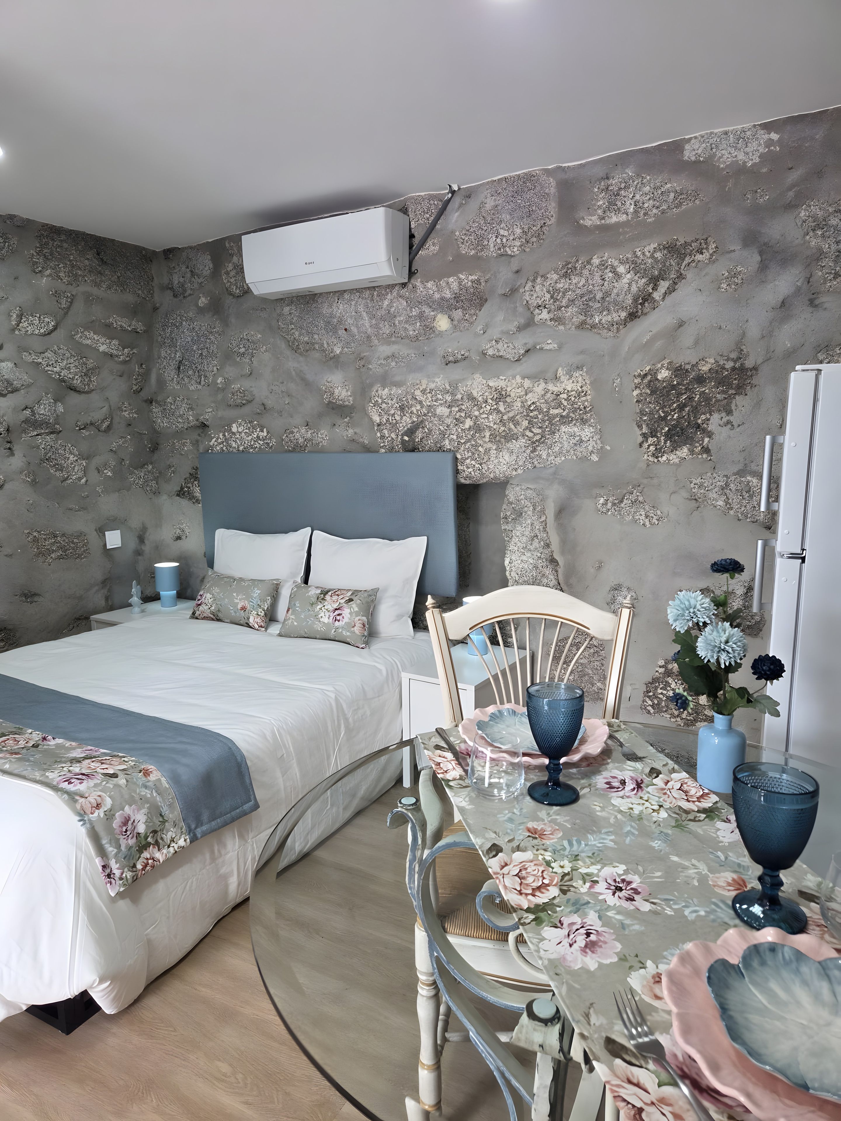 A bedroom with a stone wall, a bed with a blue headboard, a dining table with floral tablecloth, and a vase with flowers.