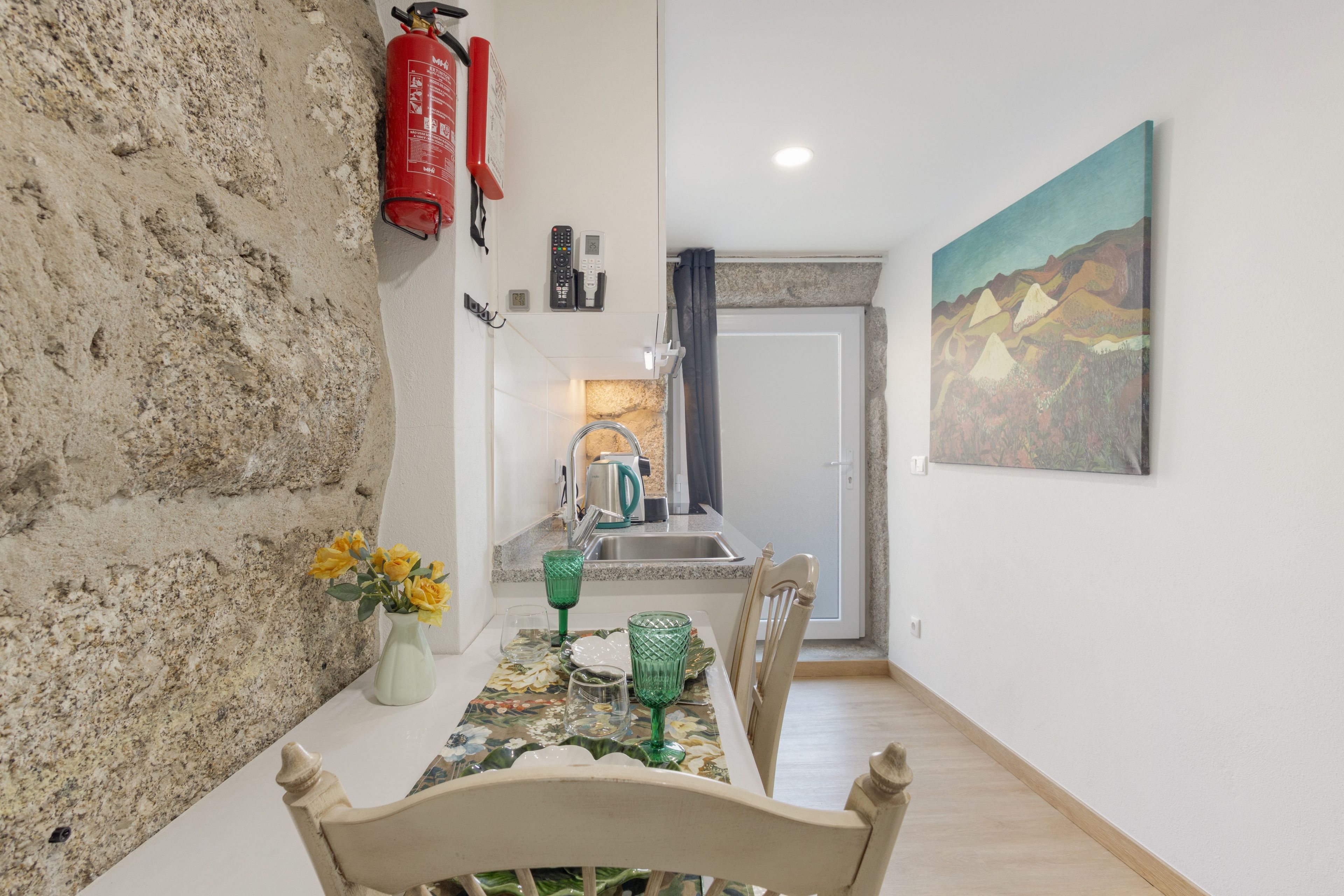 Een eethoek met een tafel gedekt voor twee personen, een keuken op de achtergrond, een brandblusser aan de muur en een schilderij.